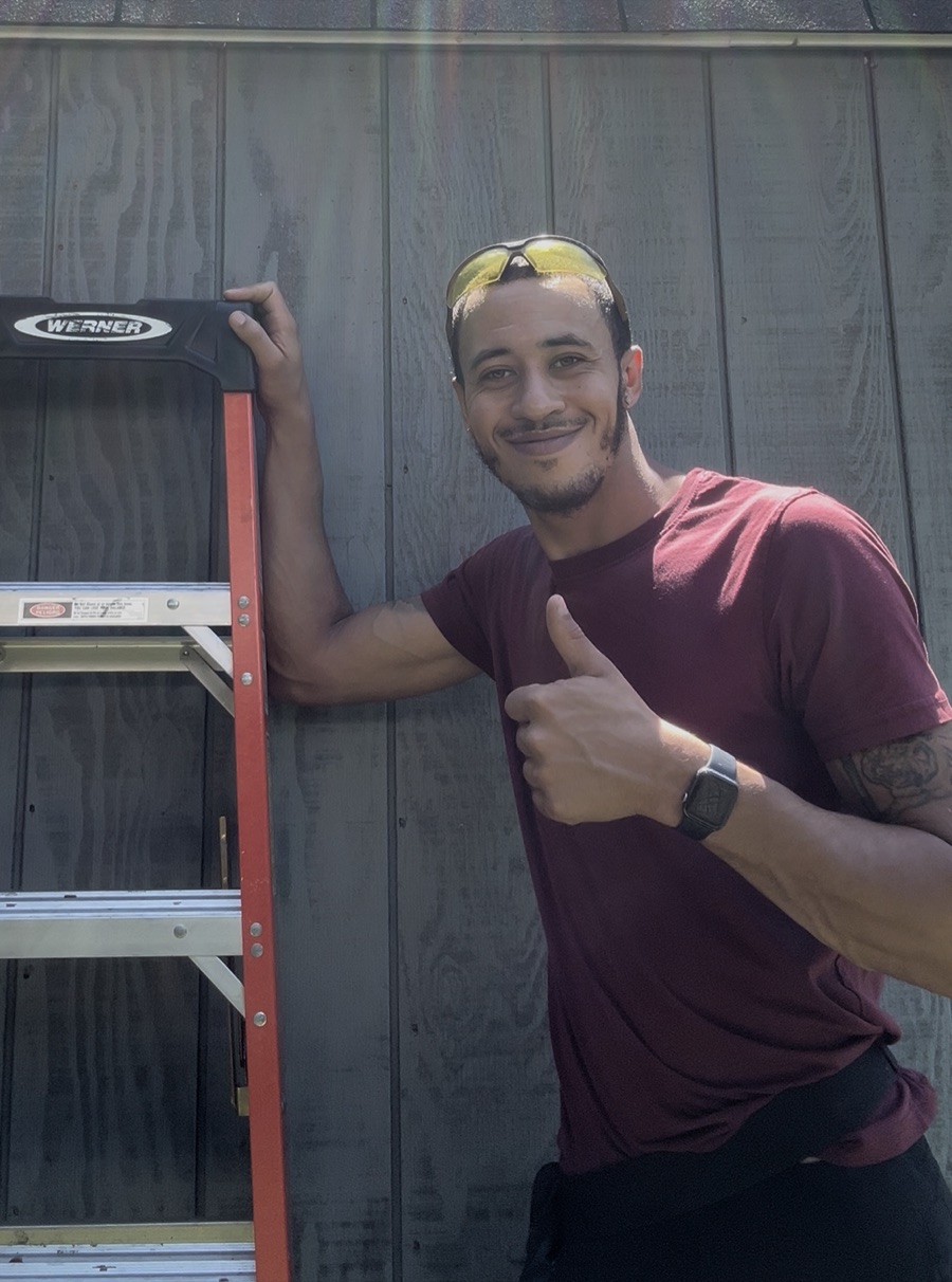 Kyle Redman standing beside a ladder against a stained wood-sided wall, giving a thumbs up